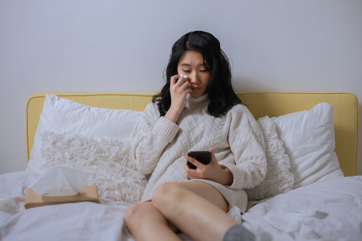 10 Harsh Truths About Why Online Dating Is Really Terrible for Your Health 2 Asian woman in a sweater, looking sad and wiping tears, holding a smartphone seated on a bed.