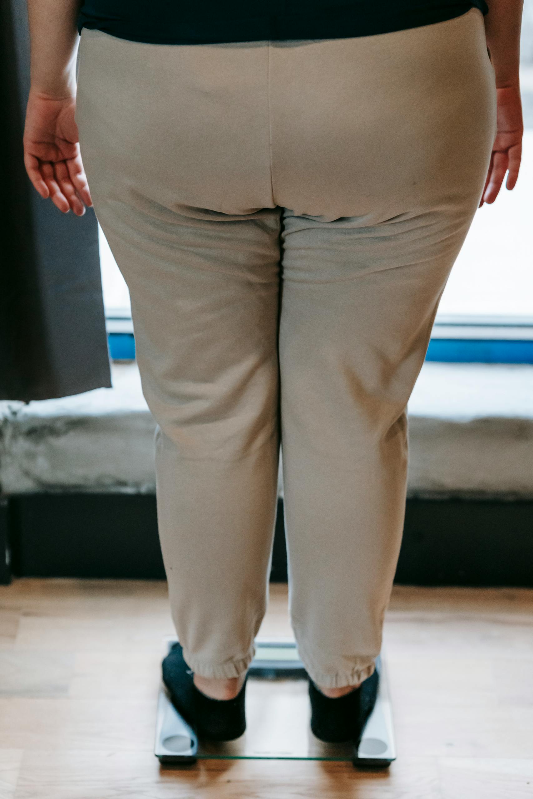 Back view of crop unrecognizable fat female in sportswear weighing on glass scale in front of window