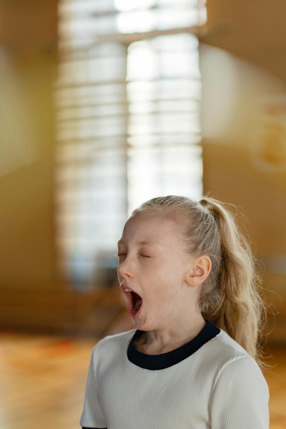 Blonde child with eyes closed yawning in a softly lit indoor setting with a blurred background.