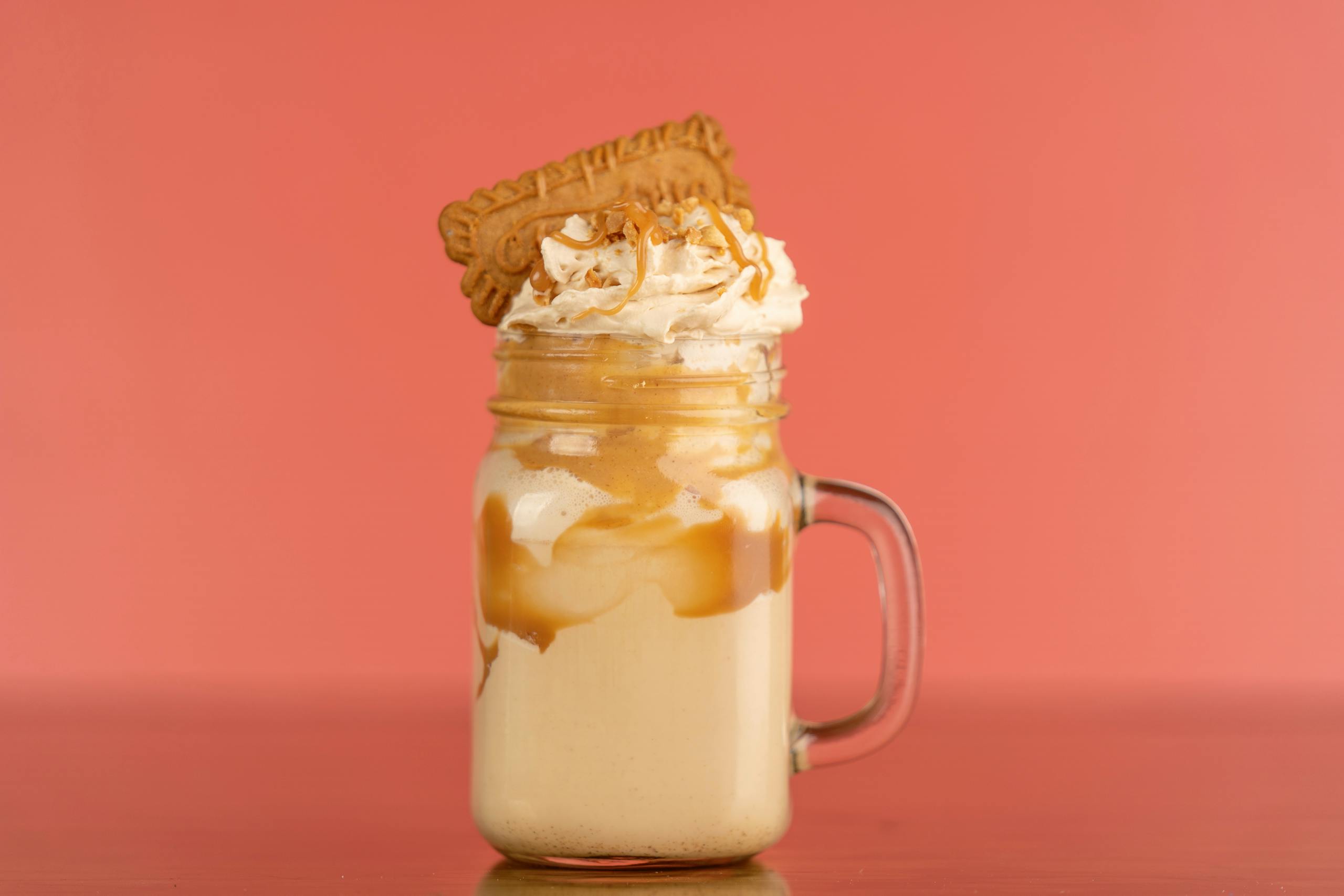 Close-up of a caramel milkshake with whipped cream and biscuit topping against a salmon background.