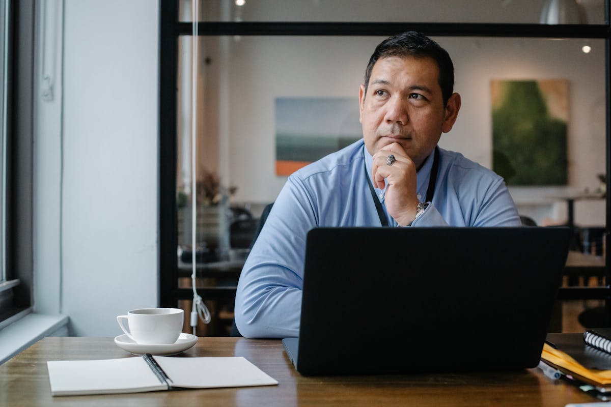 Concentrated ethnic adult male entrepreneur thinking on project while working at table with netbook and cup of drink