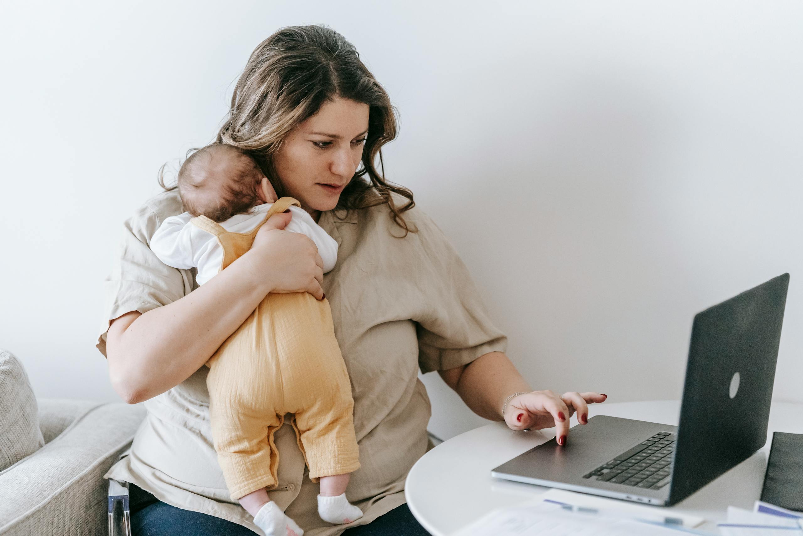 7 Uncomfortable Reasons More Women Are Saying No to Motherhood 2 Concentrated young female freelancer embracing newborn while sitting at table and working remotely on laptop at home