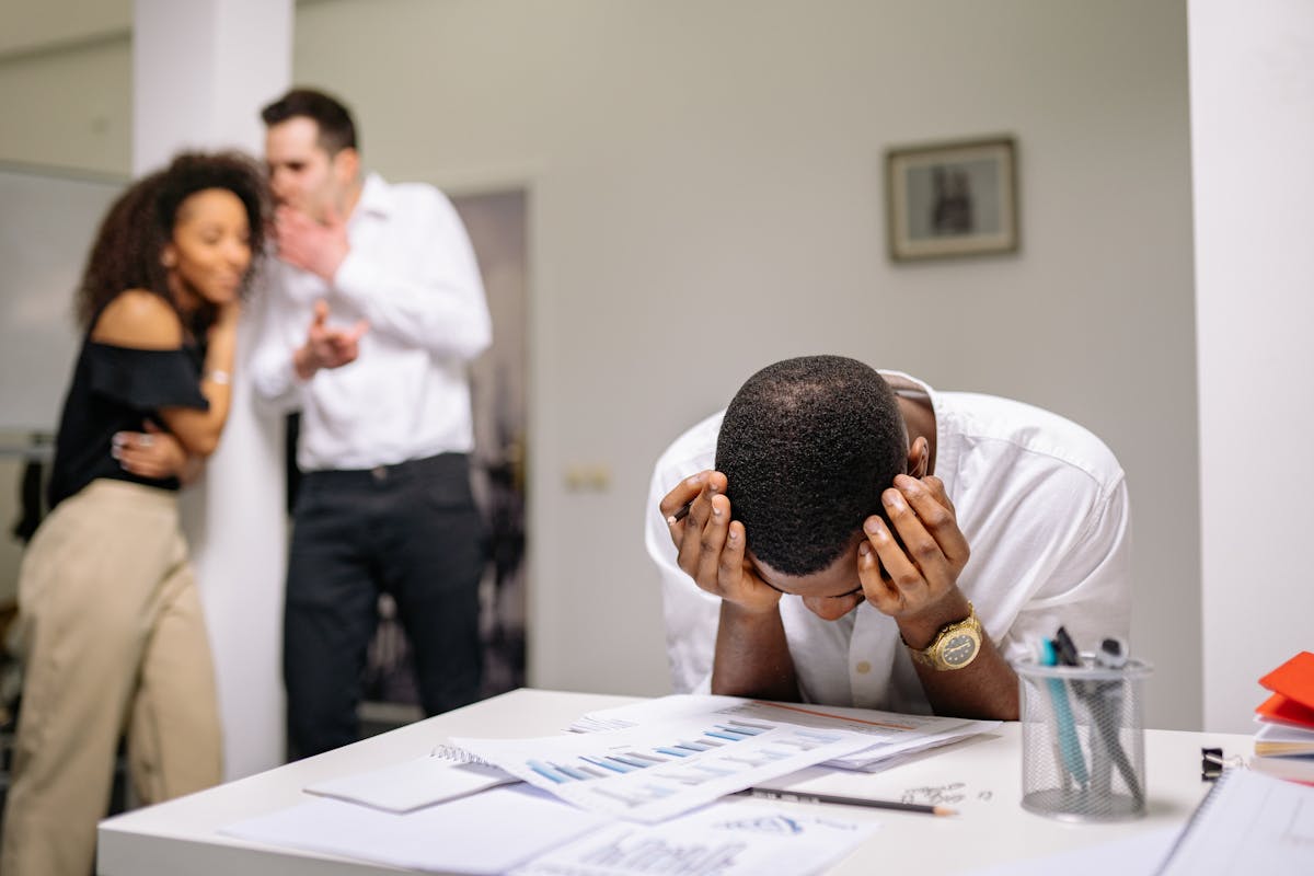 Despairing worker at office desk as colleagues gossip in background illustrating workplace bullying. Gossiping
