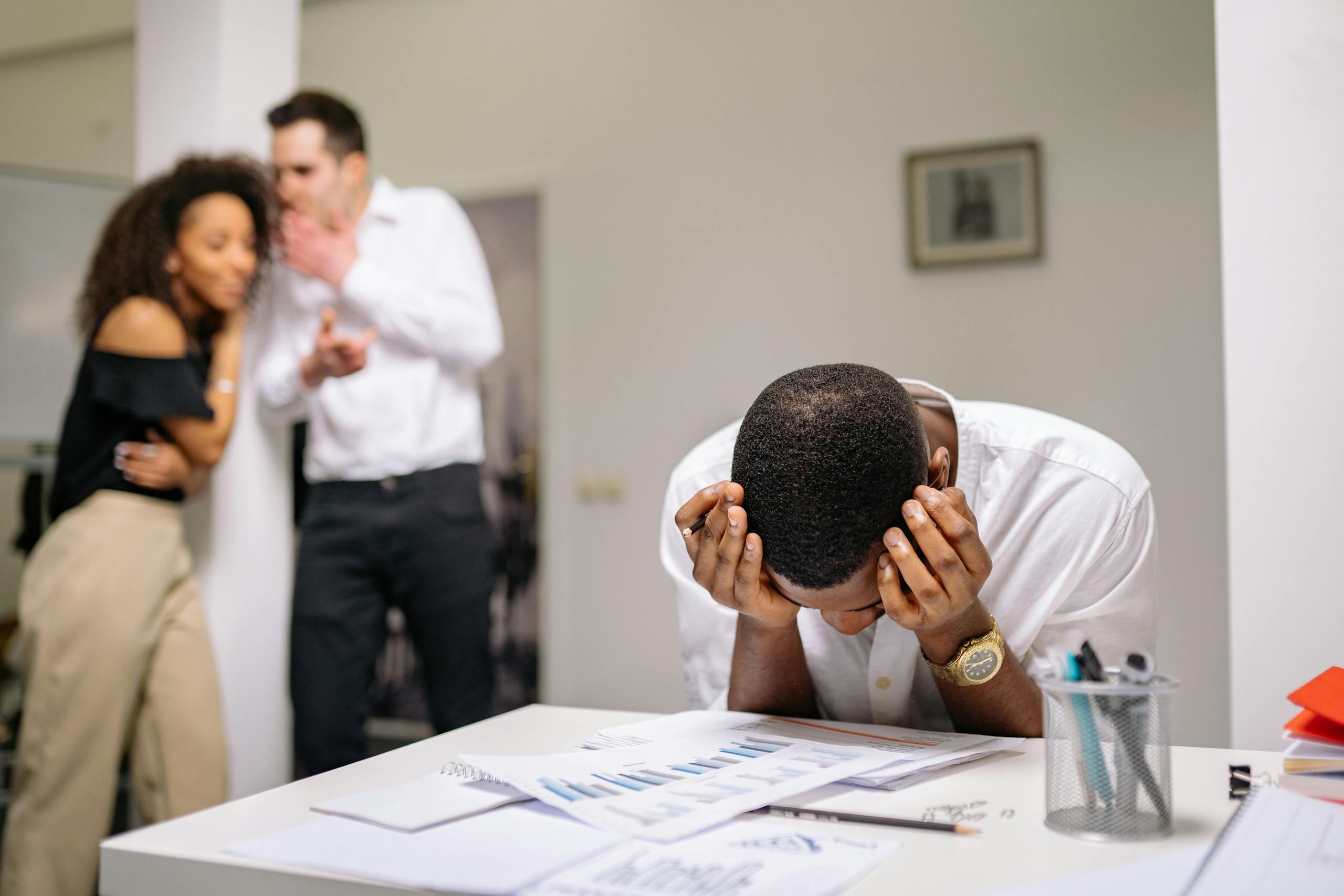 Despairing worker at office desk as colleagues gossip in background illustrating workplace bullying. Gossiping