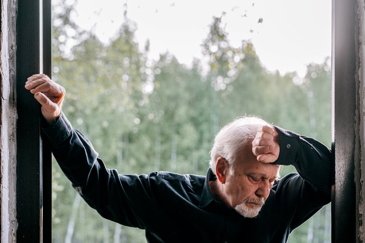 7 Disturbing Early Signs of Dementia That Have Nothing to Do With Memory Loss 4 Elderly man in black long sleeves showing emotion by an open doorway with a forest view.