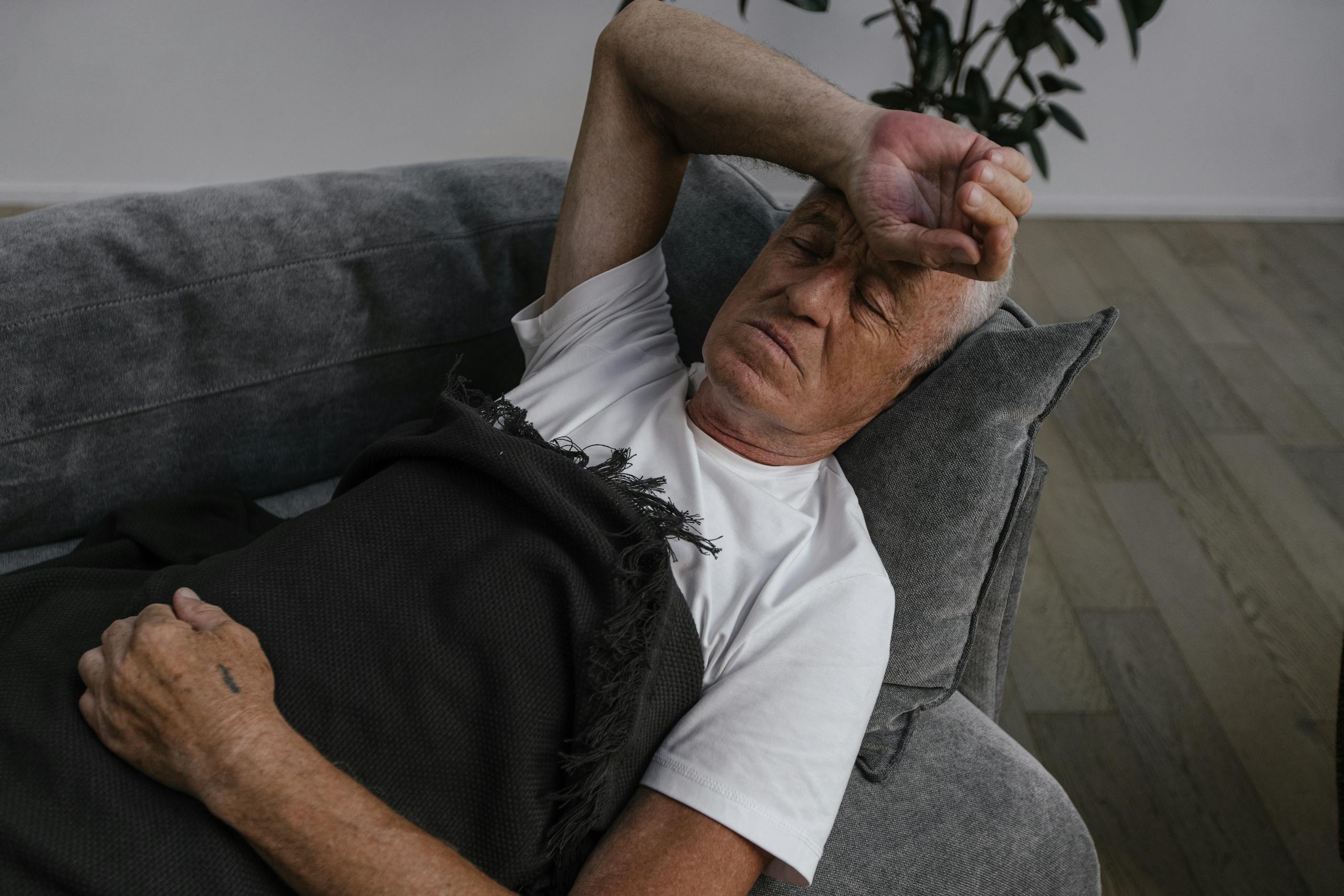 Elderly man relaxing on a sofa, hand on forehead, white shirt and blanket.