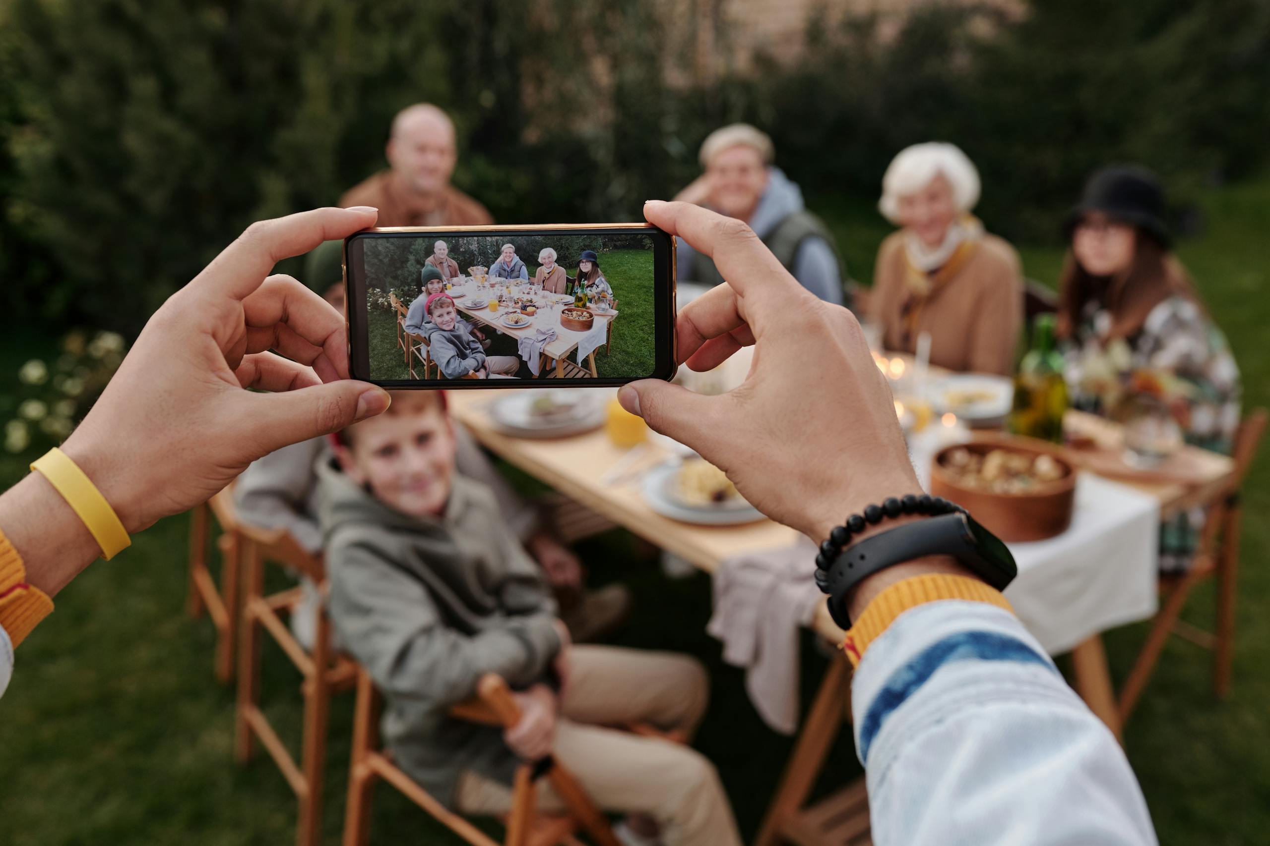 8 Things Gen Z Does That Older Americans Secretly Can’t Stand 2 Faceless person with fitness bracelet taking photo with smartphone of big multigenerational family dinner served outside
