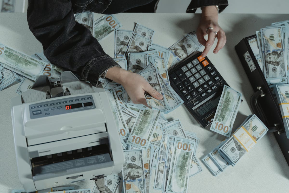 Hegseth Says Iran War Could Cost $37.5 Billion as Senators Press Pentagon on What Has Actually Been Spent. 2 Hands counting 100 dollar bills using a calculator and money counter on a table.
