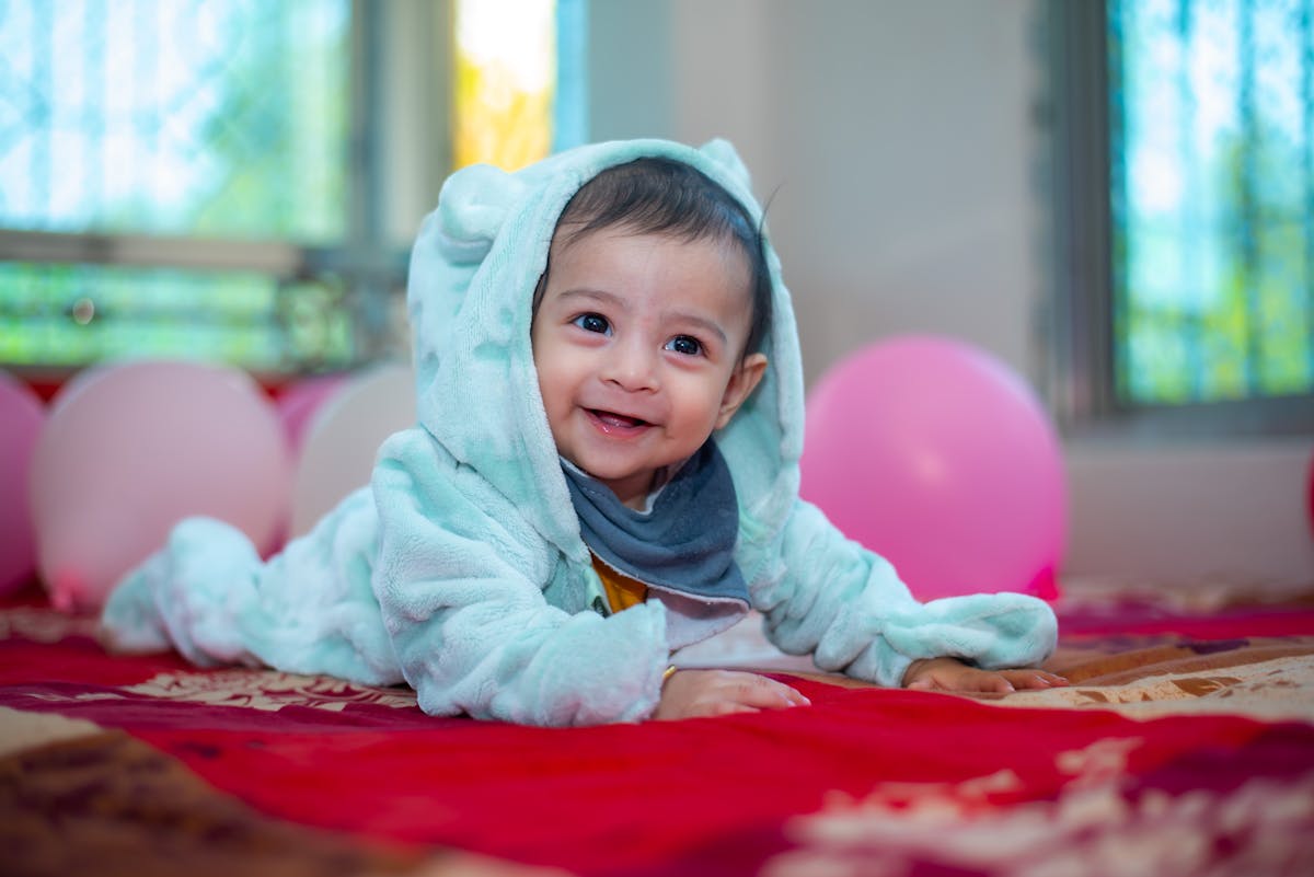 Happy baby in a hooded outfit crawling indoors surrounded by pink balloons.