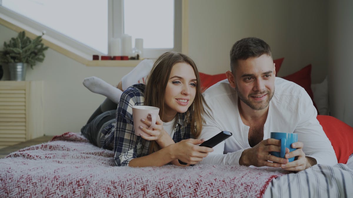 10 Tactics Narcissists Use to Hurt You Intentionally 3 Happy couple lounging on a bed, watching TV and enjoying drinks at home.