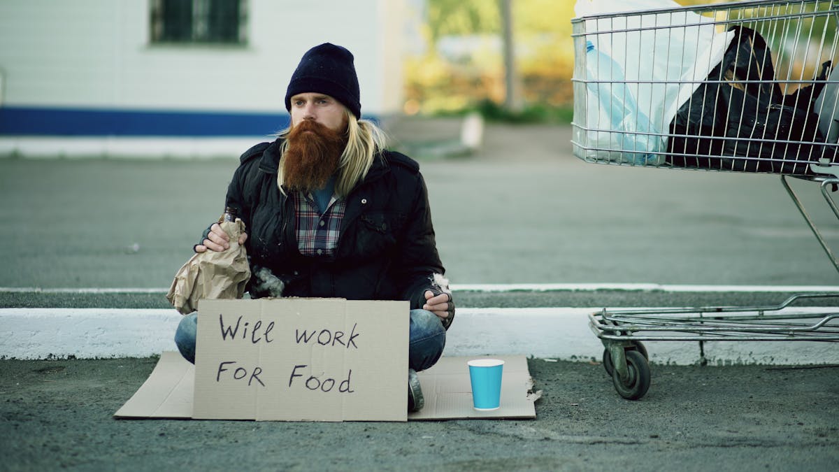 Man sitting outdoors with a sign reading 'Will work for food', next to shopping cart.