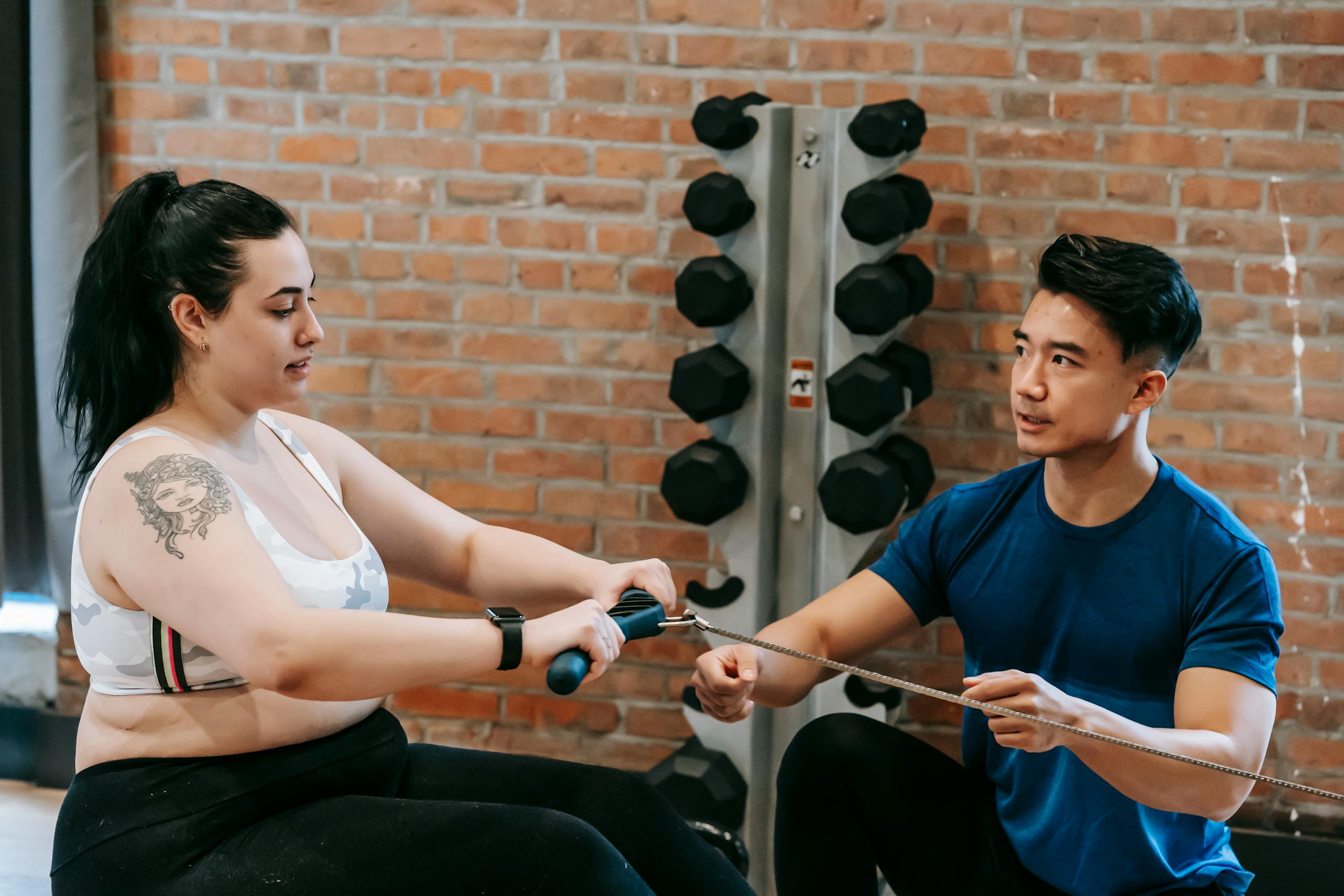 Multiethnic trainer and overweight woman doing exercises with rowing machine together in sport club