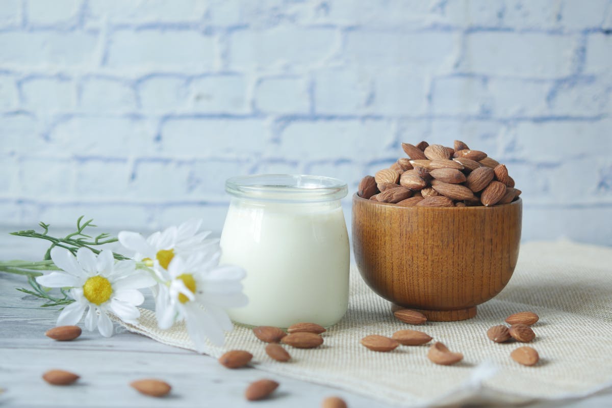 Natural almonds in a wooden bowl with a glass jar of milk and white chamomile flowers on a wood table.