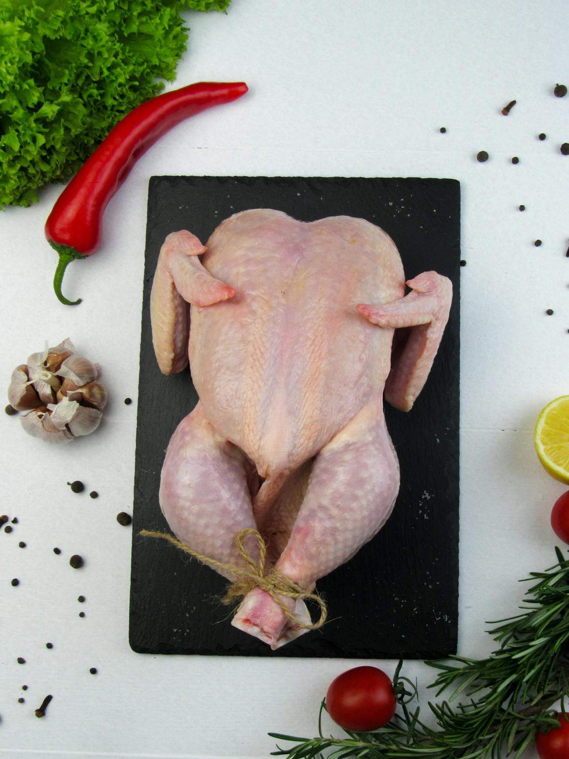 Overhead shot of a whole raw chicken surrounded by fresh spices and vegetables on a slate board.