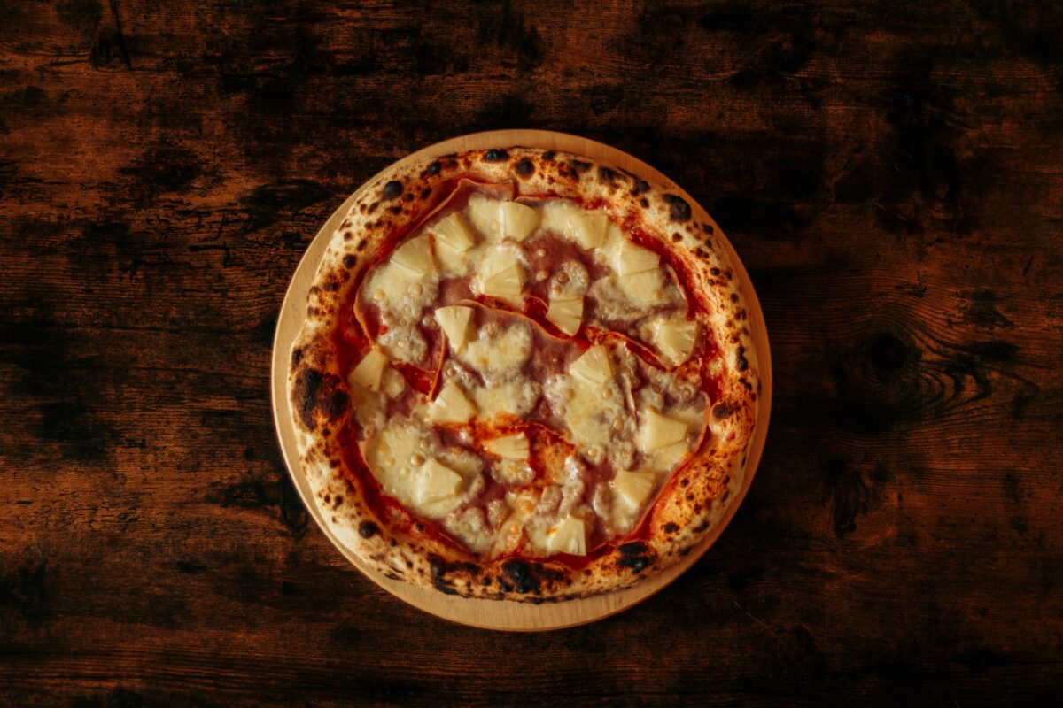 10 Famous Dishes with Misleading Names 1 Top view of a Hawaiian pizza with pineapple and ham on a rustic wooden table.