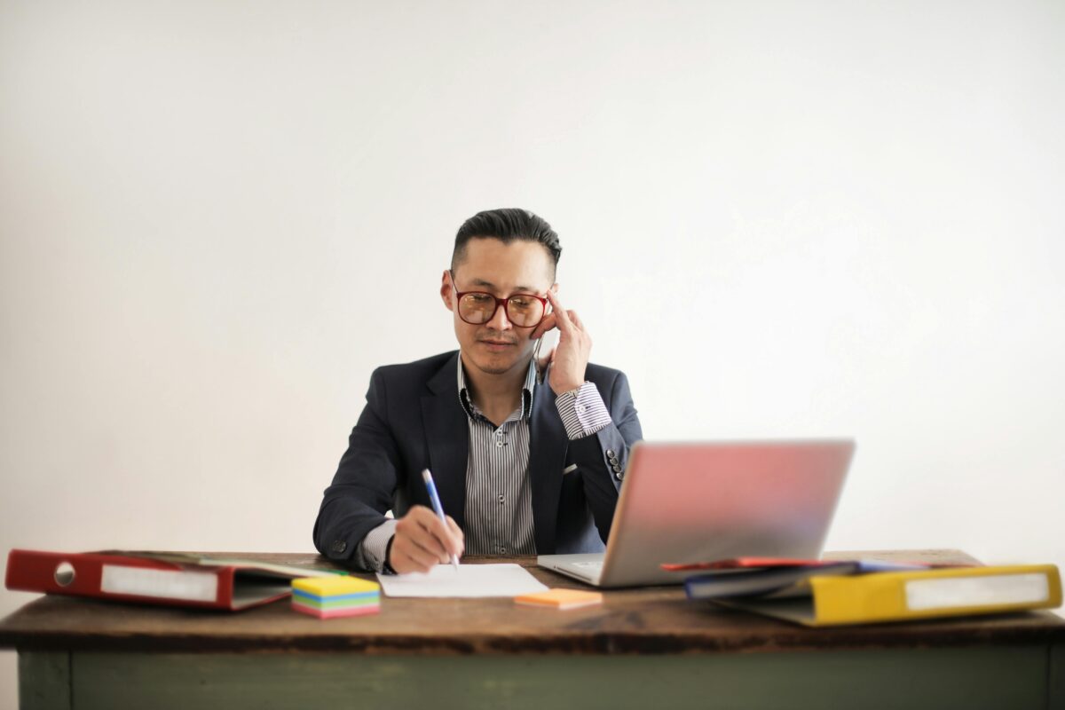 photo-of-man-in-blue-suit-jacket-striped-shirt-and-eyeglasses-talking-on-the-phone-while-sitting-at-a-table-with-his-laptop