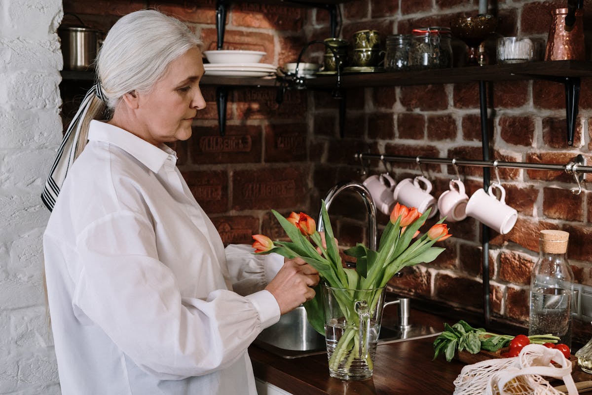 Senior woman in a white shirt arranging tulips in a cozy kitchen with brick walls.