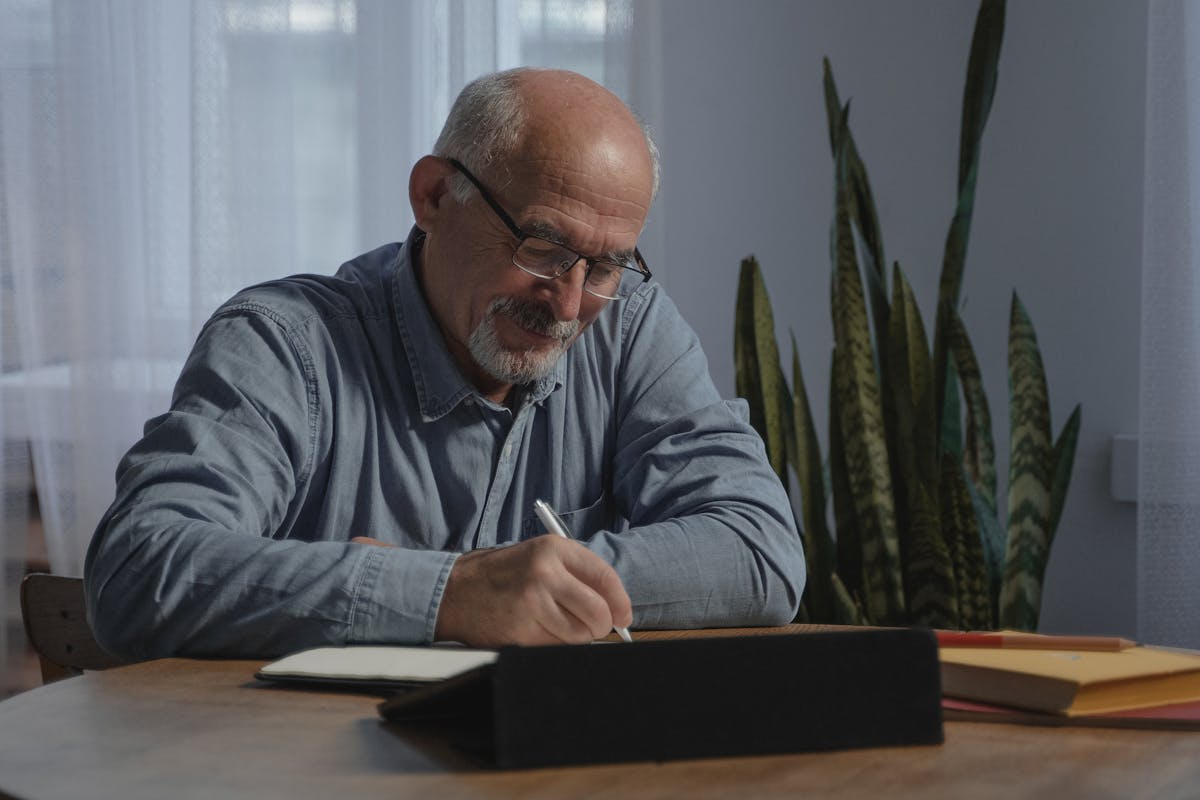50 Isn’t Too Late, But These 8 Mistakes Make It Harder 4 Smiling senior man taking notes at home, embodying focus and remote work lifestyle.