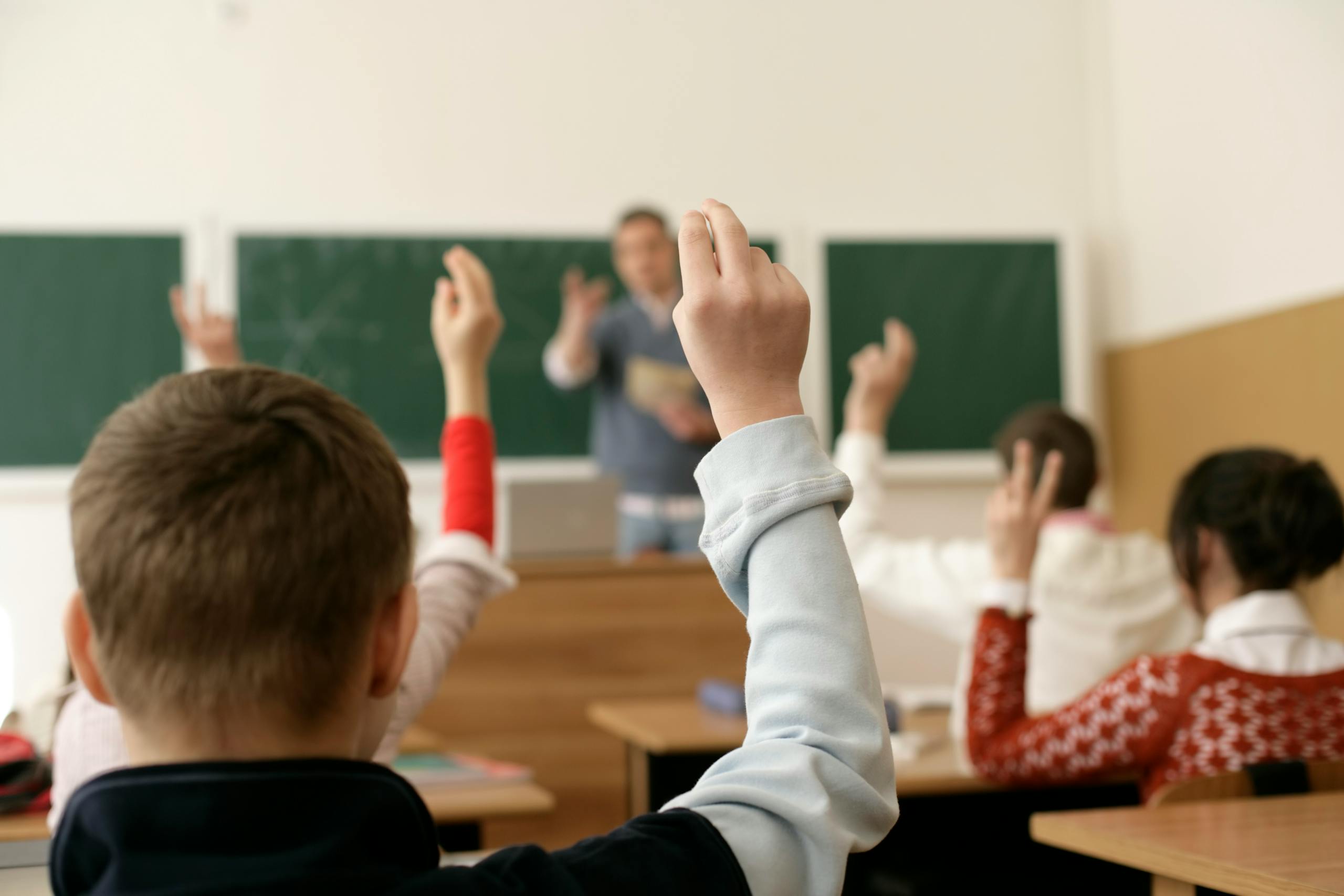 10 Outrageous Ways Your Local School Is Quietly Brainwashing Your Kids 1 Students in a classroom raising hands, engaging in a lesson with the teacher.