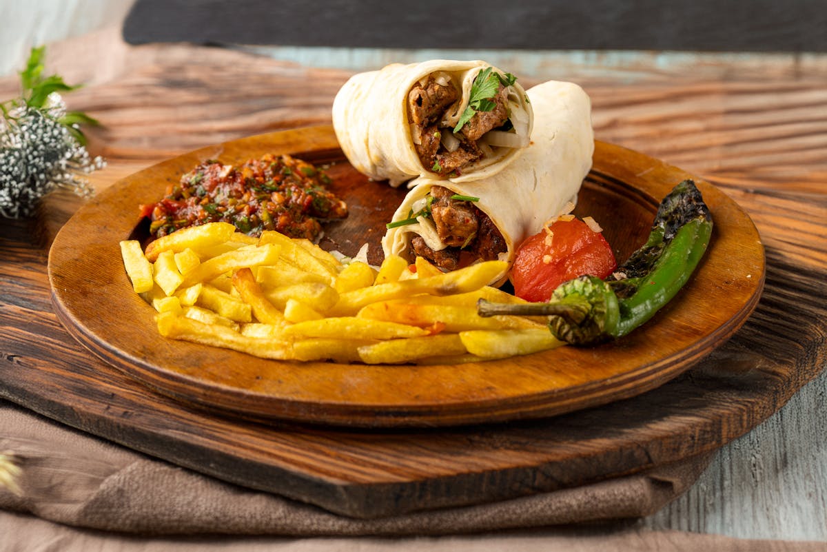 Tasty shawarma wrap on a wooden plate with fries, salsa, and grilled vegetables.