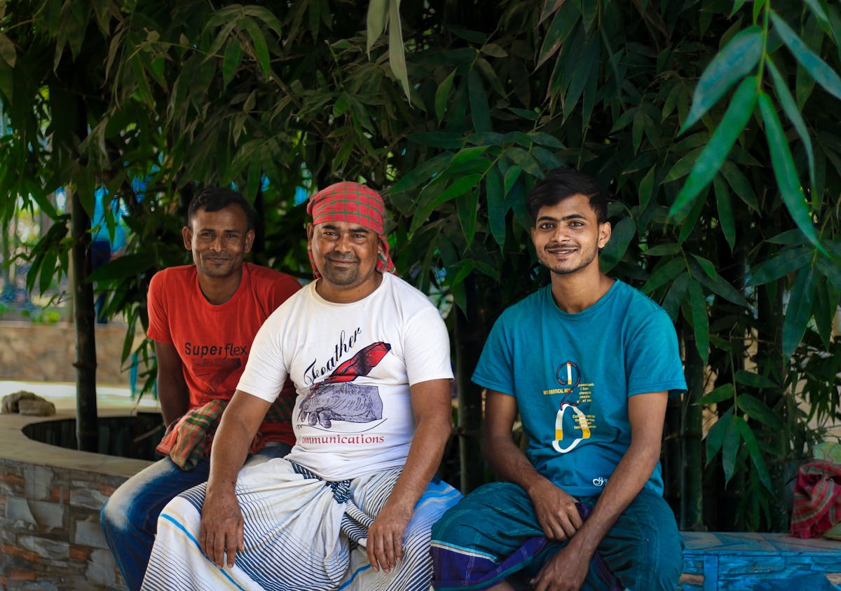 Three men sitting together, smiling, in an outdoor setting with bamboo plants in Dhaka.