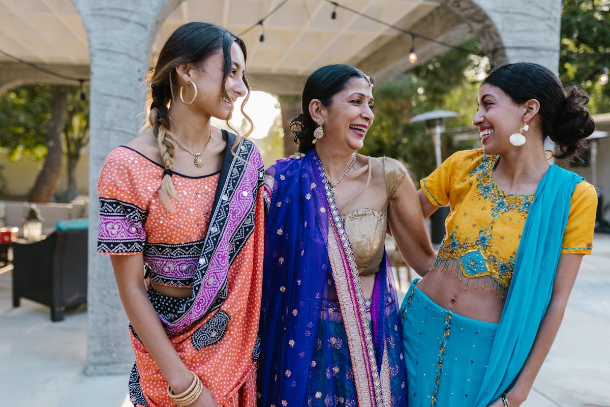 8 Countries Where People Wear Their Underwear the Longest 3 Three women smiling and embracing in colorful Indian saris, celebrating a joyous occasion outdoors.