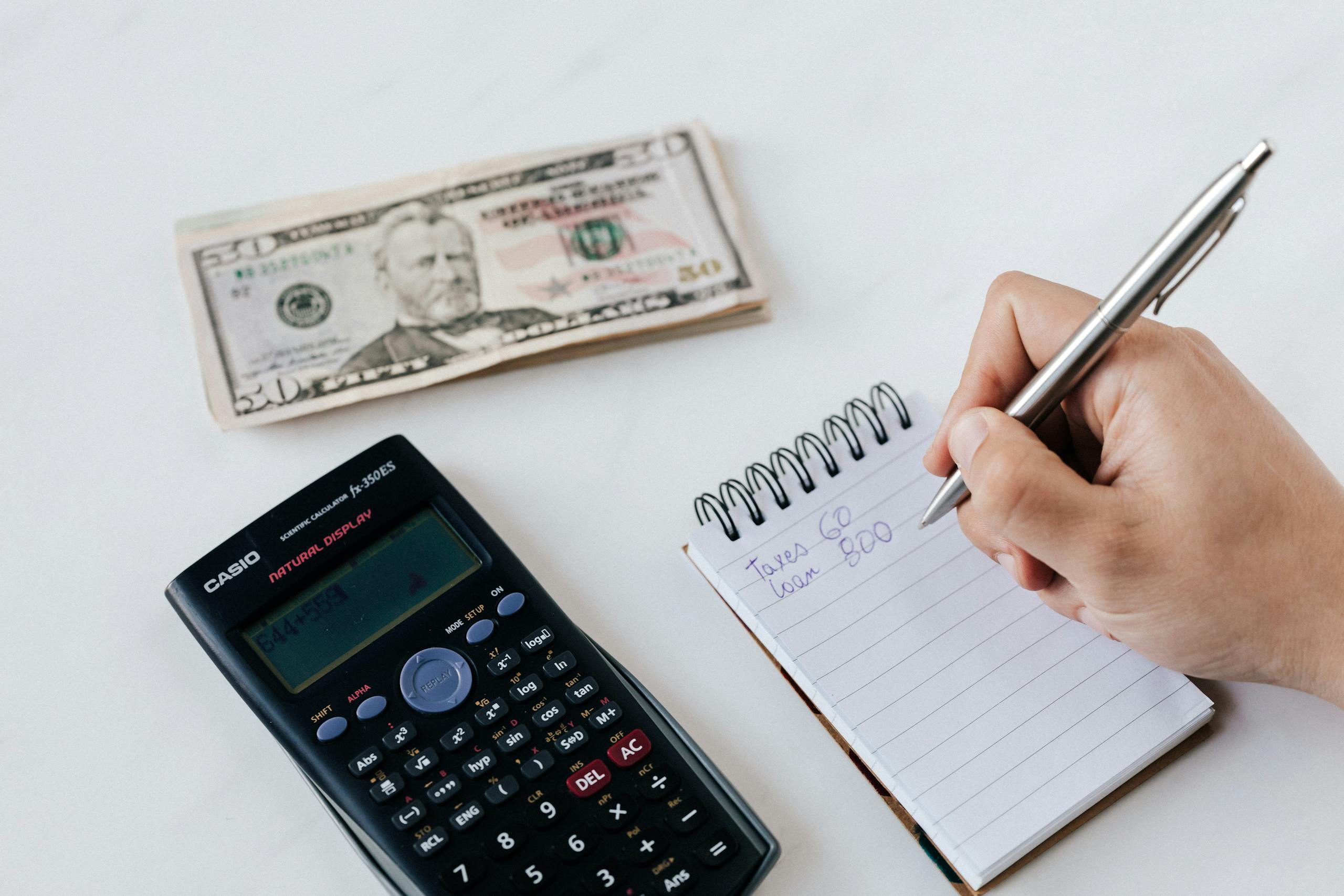 9 Brutal Ways Student Loans Are Still Controlling American Adults 4 Top view of crop faceless accountant using pen and notepad while counting expenses on table near pile of paper money and calculator