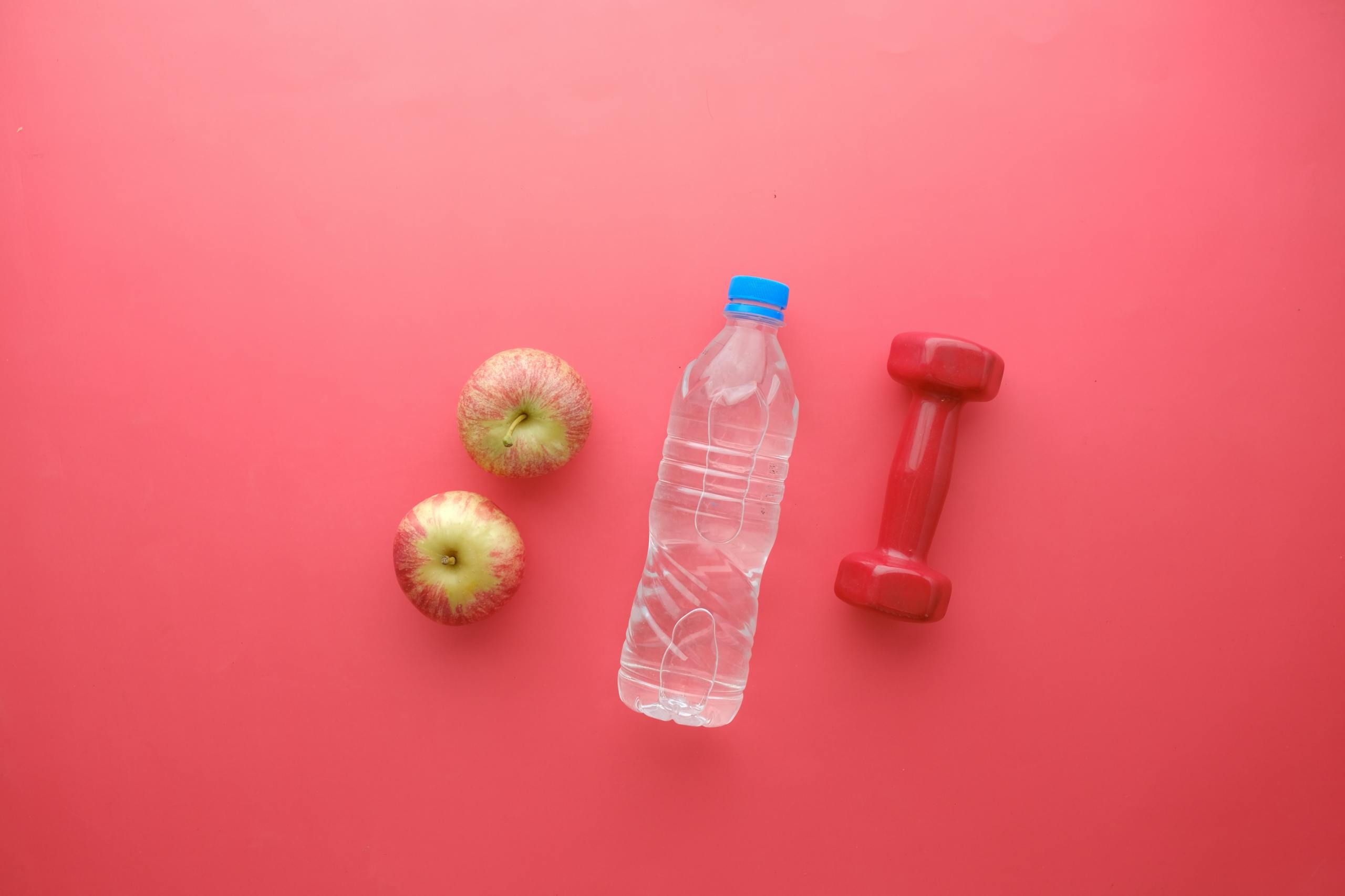 Top view of water bottle, apples, and dumbbell on bright red background. Perfect for fitness and wellness themes.