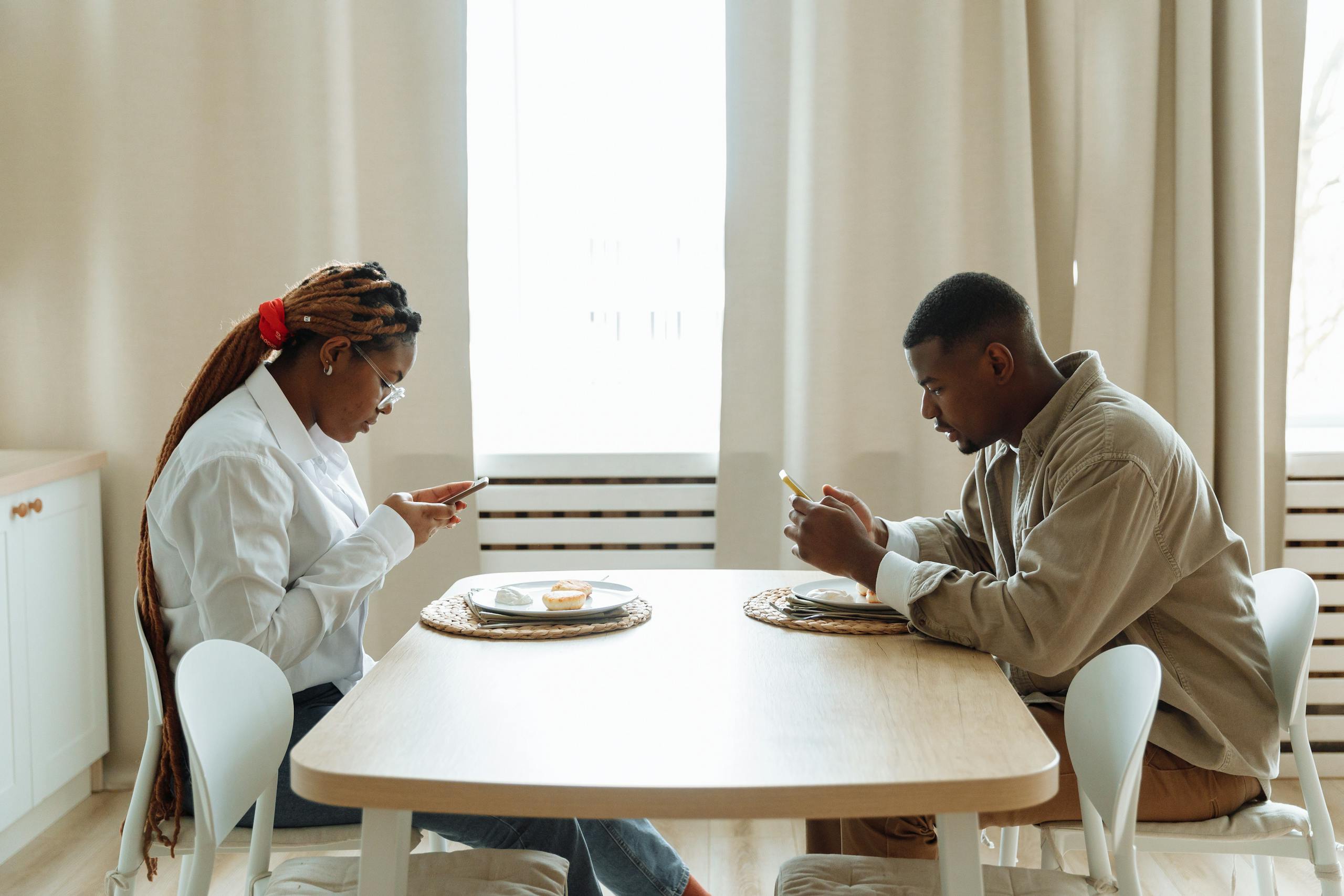 8 Ugly Reasons Social Media Is Destroying Attention Spans 4 Two adults sitting at a dining table, focused on their smartphones while having breakfast indoors.