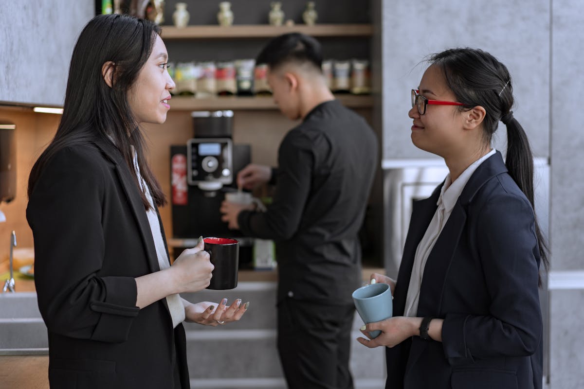 Two female colleagues engaging in conversation during a coffee break in a modern office setting.