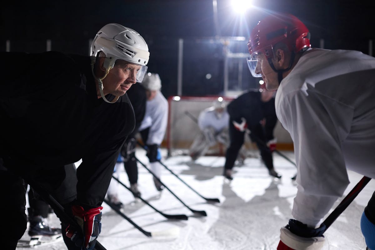 50 Isn’t Too Late, But These 8 Mistakes Make It Harder 2 Two ice hockey players faceoff on a rink during a night game, surrounded by teammates in action.