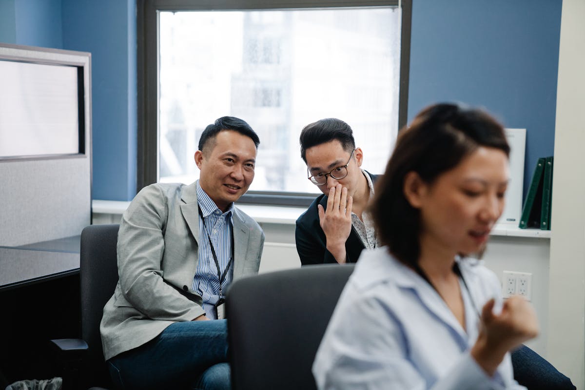 Two male colleagues sharing a secret in an office while a female coworker works nearby.