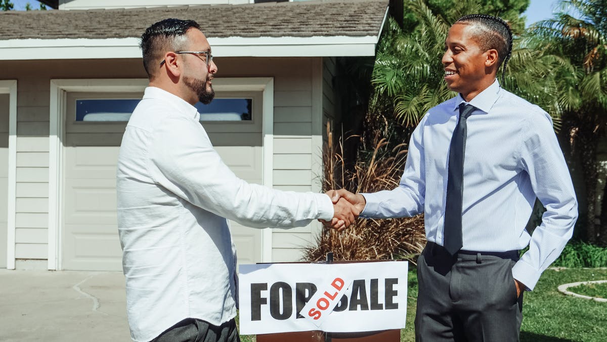 9 beliefs Gen X still holds that no longer apply today 3 Two men shaking hands in front of house sold sign, sealing real estate deal outdoors.