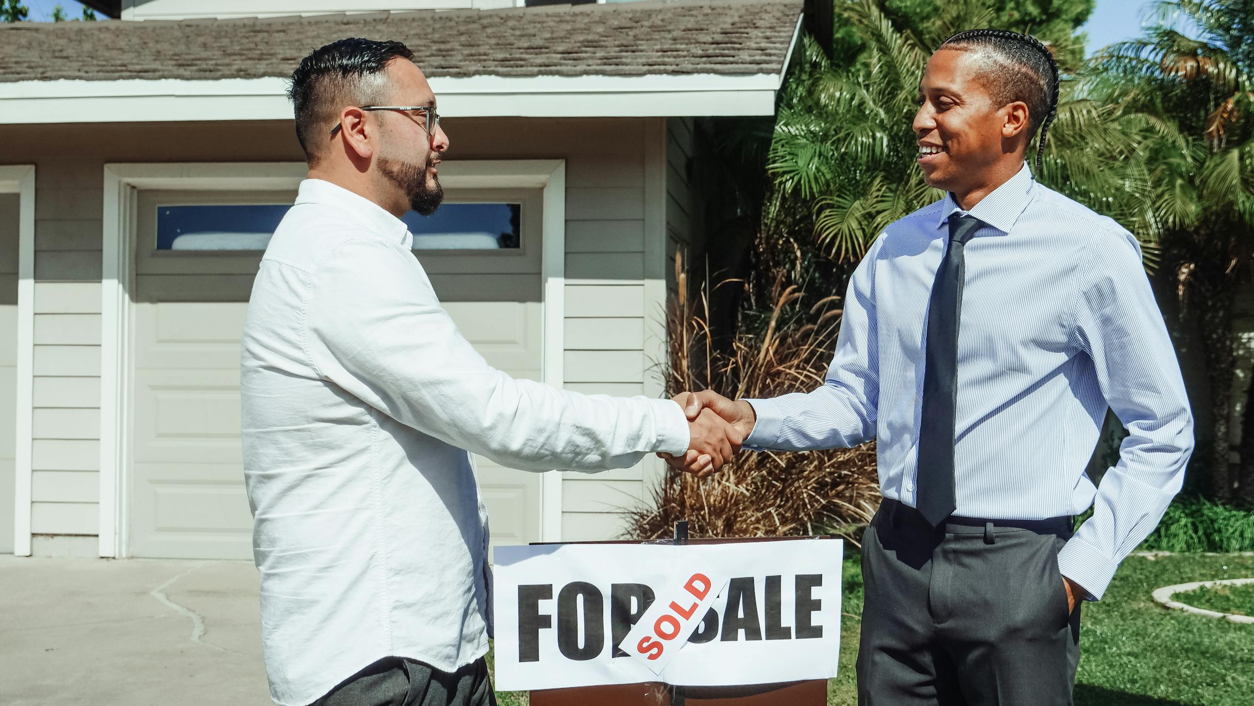 7 Reasons Americans Don’t Believe the Economy Is Fine Anymore 2 Two men shaking hands in front of house sold sign, sealing real estate deal outdoors.
