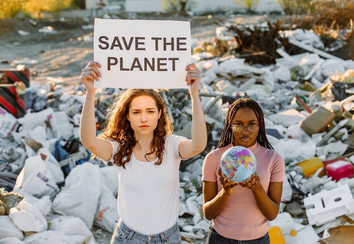 Two women demonstrate at a dumpsite, holding a sign urging to save the planet.