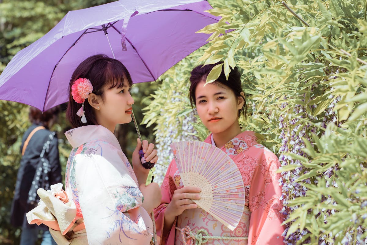 Two women in traditional kimono with umbrella and hand fan amid lush greenery and flowers.