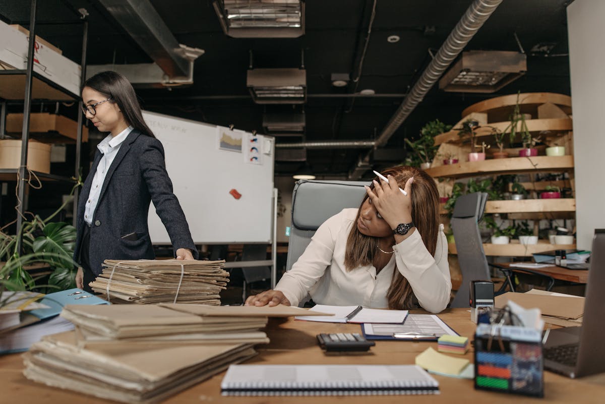 Two women working with documents in a modern office setting, one sitting at the desk, the other standing. Missing Deadline