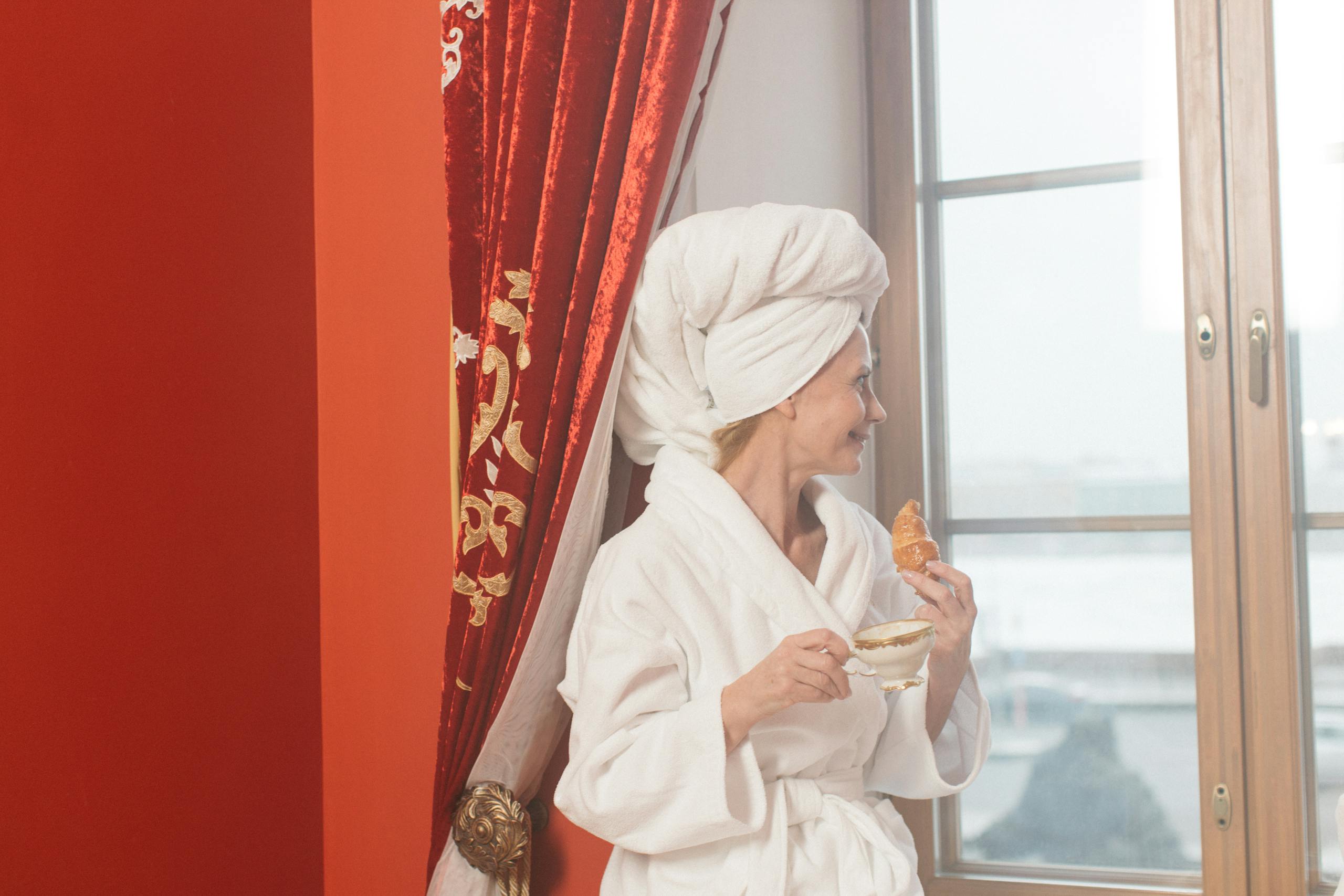 8 Hotel Mistakes That Can Ruin Your Stay 5 Woman in bathrobe with coffee and croissant looks out window, enjoying a leisurely morning.