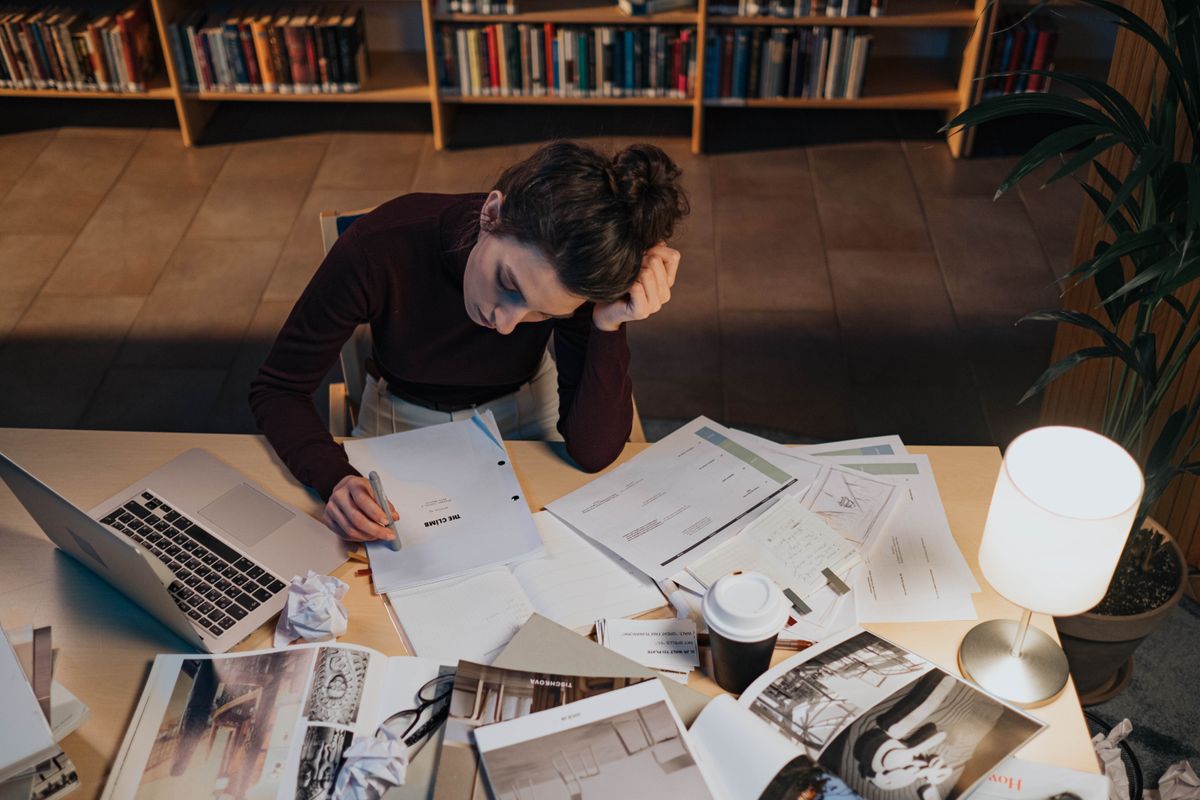 Woman studying at desk with documents, laptop, and coffee in a cozy library setting.
