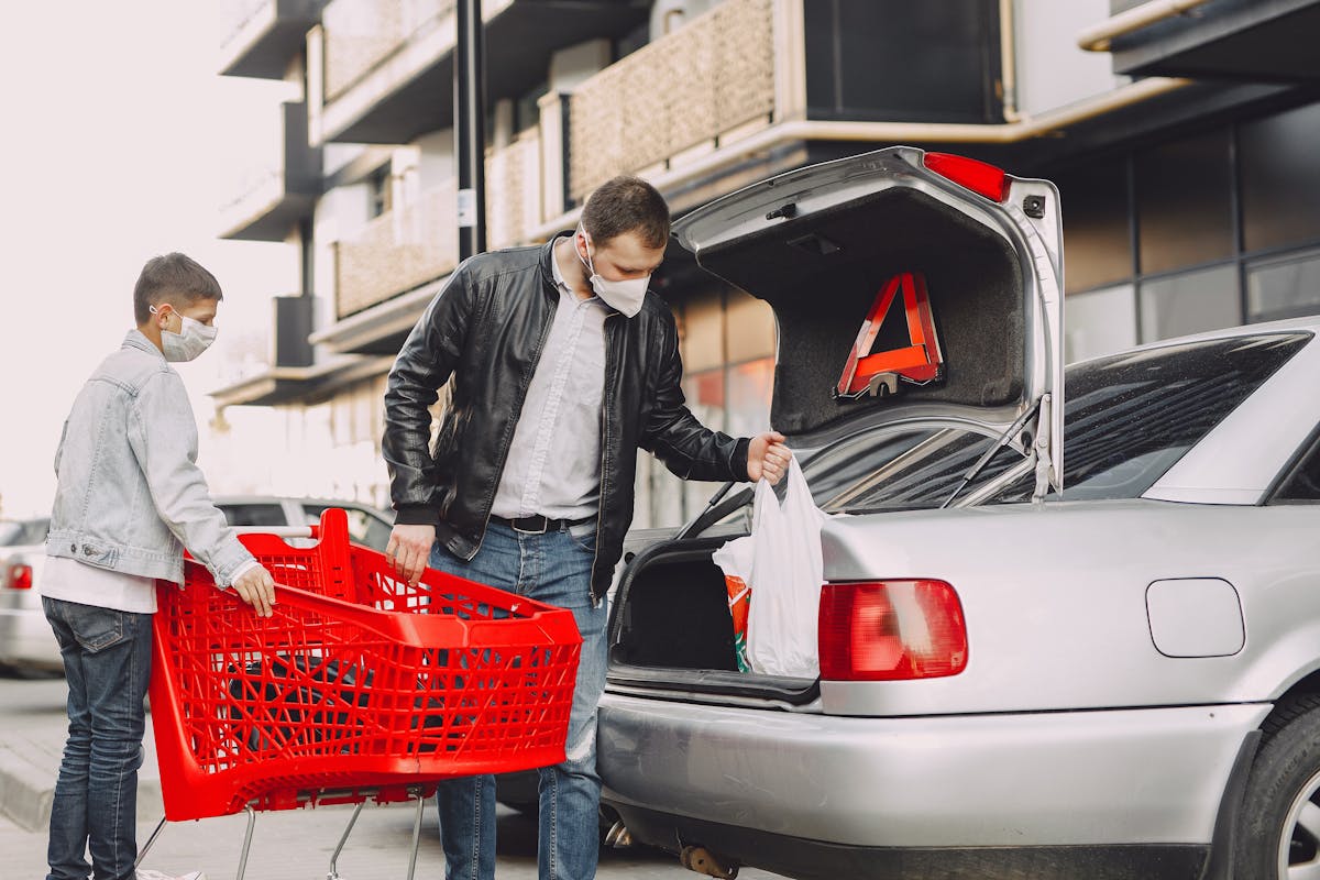 Young father and little son in casual clothes and protective masks moving shopping bags from supermarket trolley to trunk modern building parking