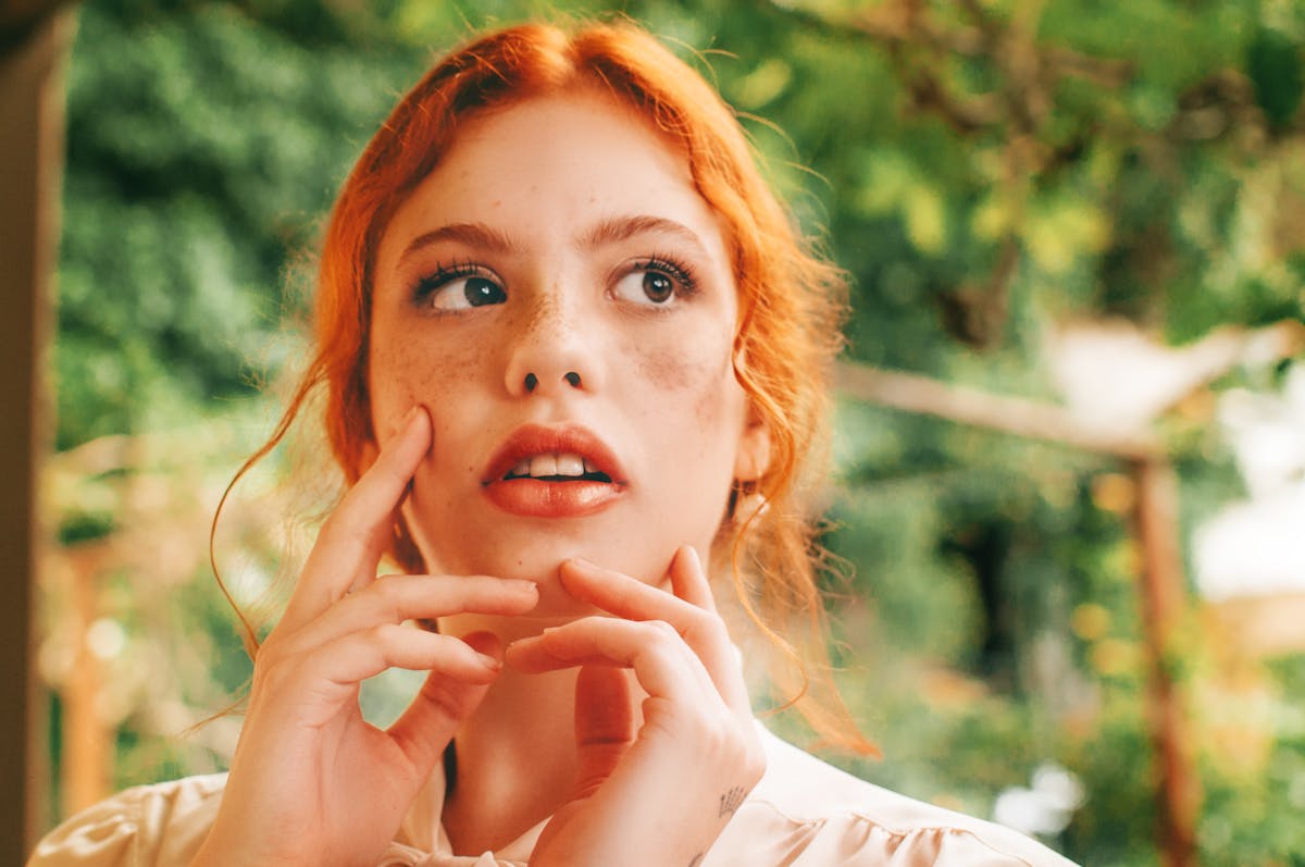 Debunking Redhead Myths: Truths About Gingers You Should Know 3 Young female with red hair with hands on face looking away thoughtfully while standing on terrace with green trees on blurred background in summer