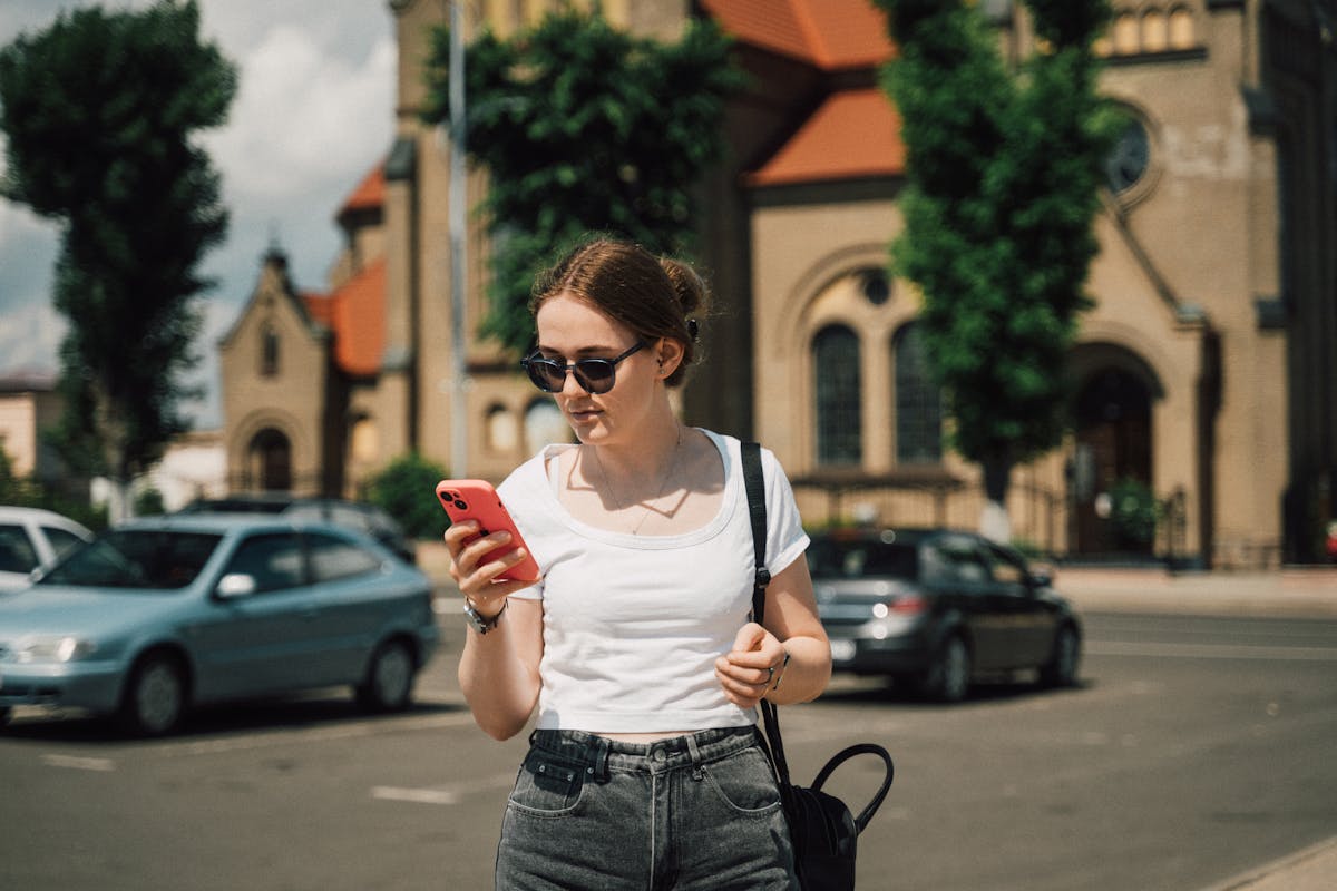 Young woman with sunglasses using smartphone in sunny urban street with historic architecture.