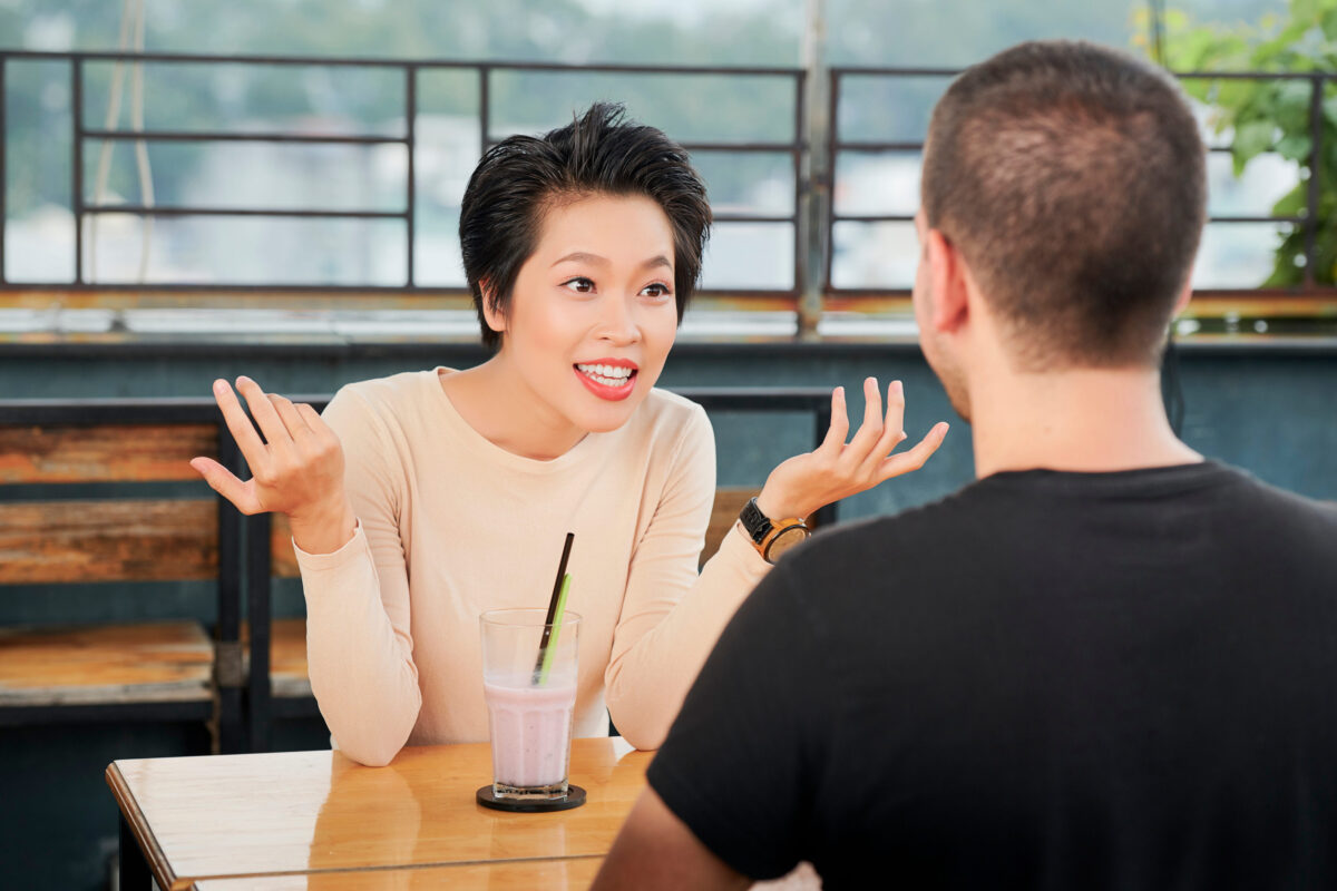 Excited Asian woman telling something to the young man while they sitting at the table and drinking juice in outdoor cafe