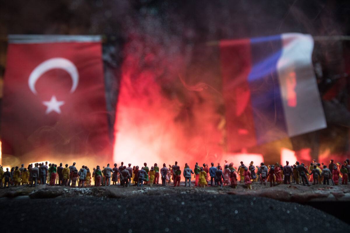 Closeup, flags of Russia and Turkey against destroyed city at night. Decorated diorama. Crowd looking on burning buildings. Concept of crisis of war. Selective focus