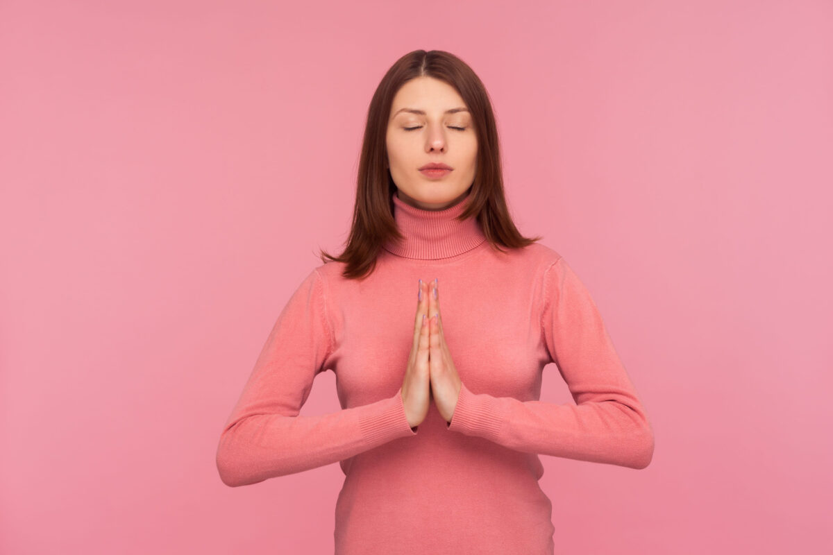 13 Things You Should Never Say to Someone With Anxiety 1 Peaceful concentrated brunette woman in pink sweater pressing hands together showing namaste gesture, practicing yoga to calm down. Indoor studio shot isolated on pink background