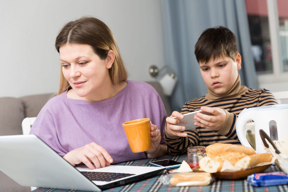 Parents Are on Devices During Meals Even More Than Their Kids, and The Family Dinner Table is Paying The Price 2 Young woman working at laptop and son using phone at table with tea