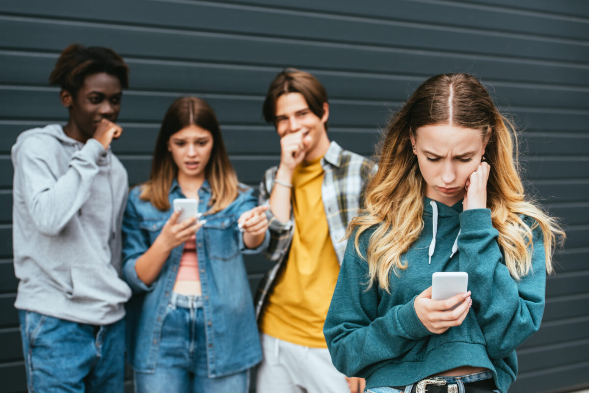 Selective focus of upset girl using smartphone near laughing multiethnic teenagers outdoor