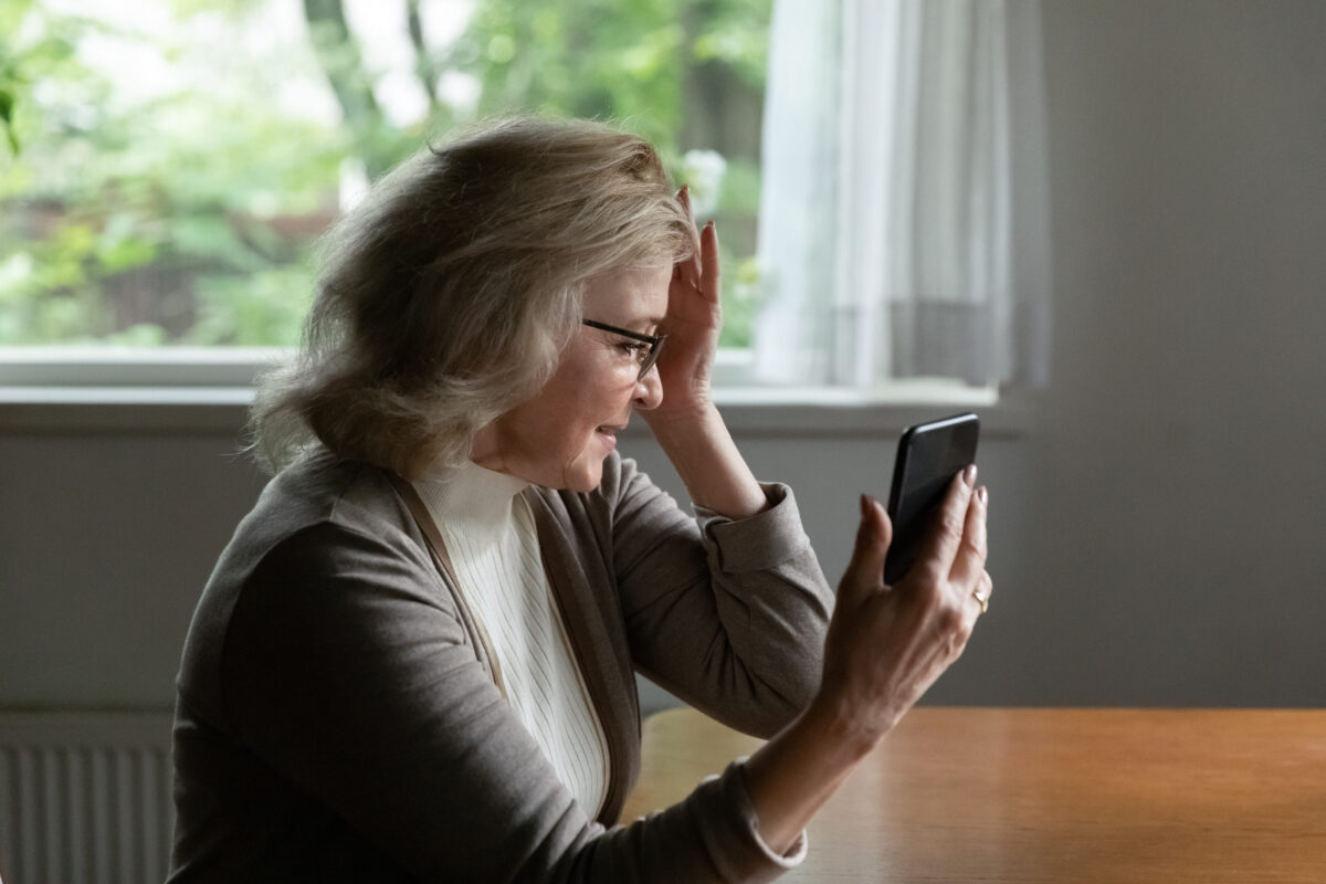 Confused senior 60s woman holding head, feeling dizziness, headache, using smartphone for video call to doctor. Pensioner reading message on mobile phone, shocked with news, suffering from memory loss