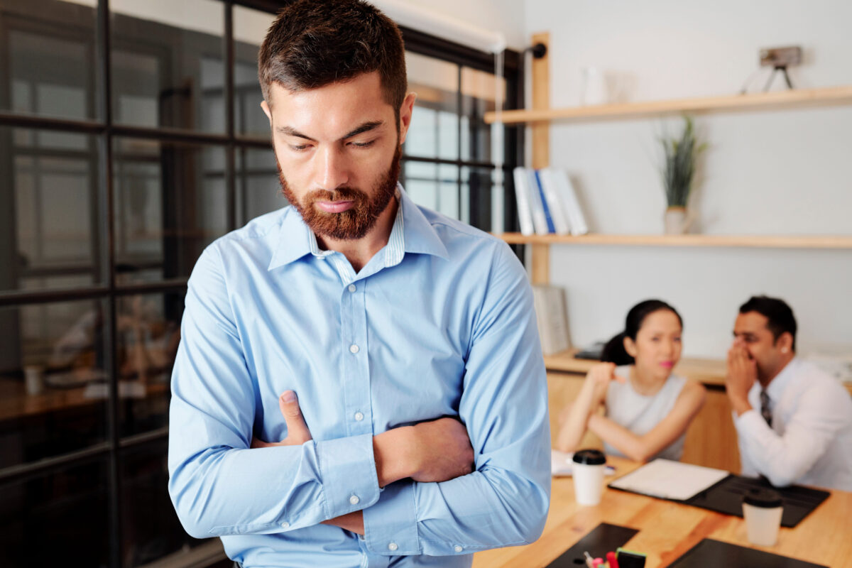 7 Worst Signs of a Weak Man Women Secretly Hate 1 Upset young businessman standing in closed pose, colleagues are gossiping about him in background