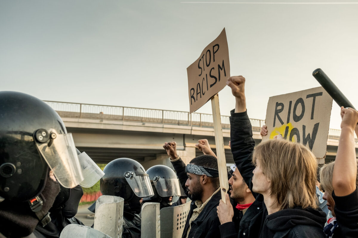 Group of displeased interracial people with signs standing in front of riot police and fighting for freedom of all ethnicities outdoors